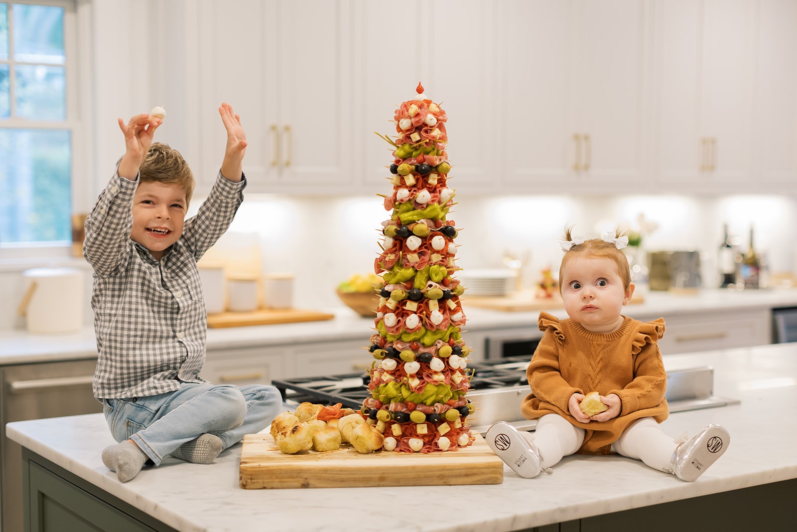 Two little kids cheering on a kitchen counter next to an antipasto tree